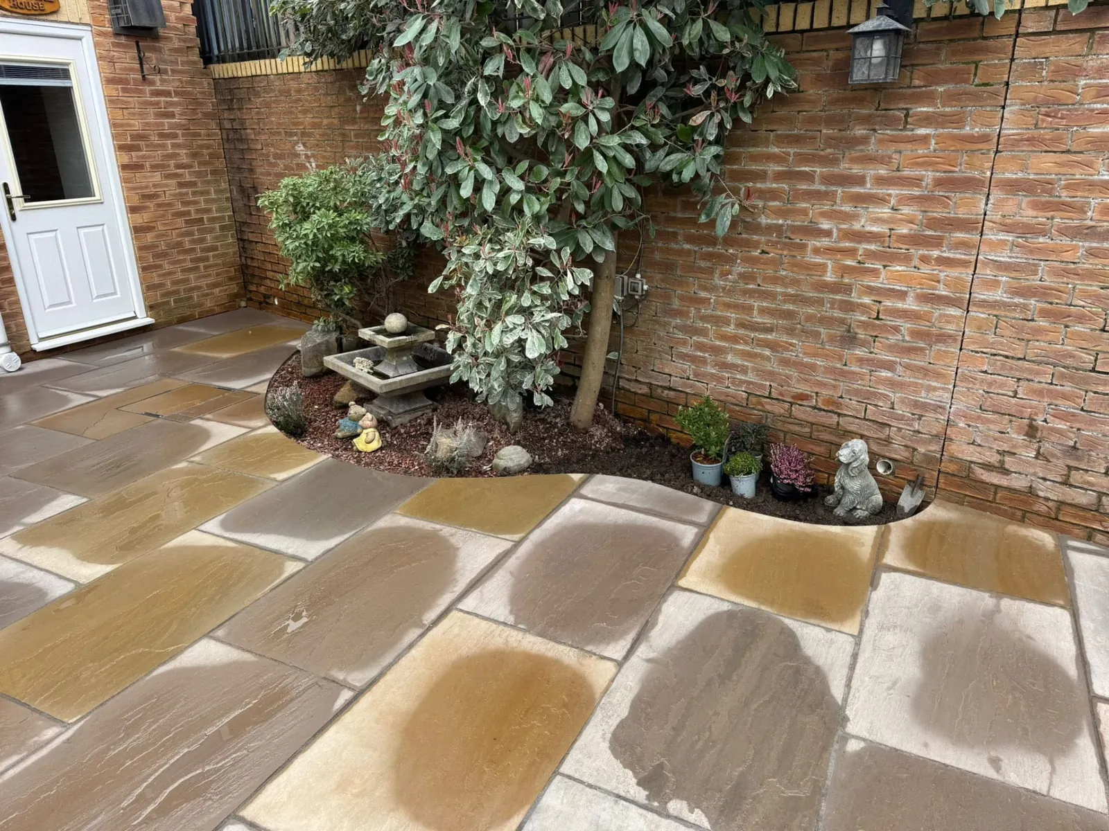 Sandstone patio laid around a planted bed beside a brick house