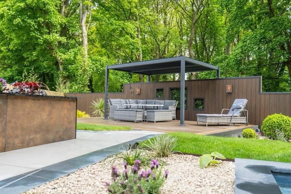 Woodland-edge garden with louvred pergola, outdoor sofa, corten planter and pale porcelain paving