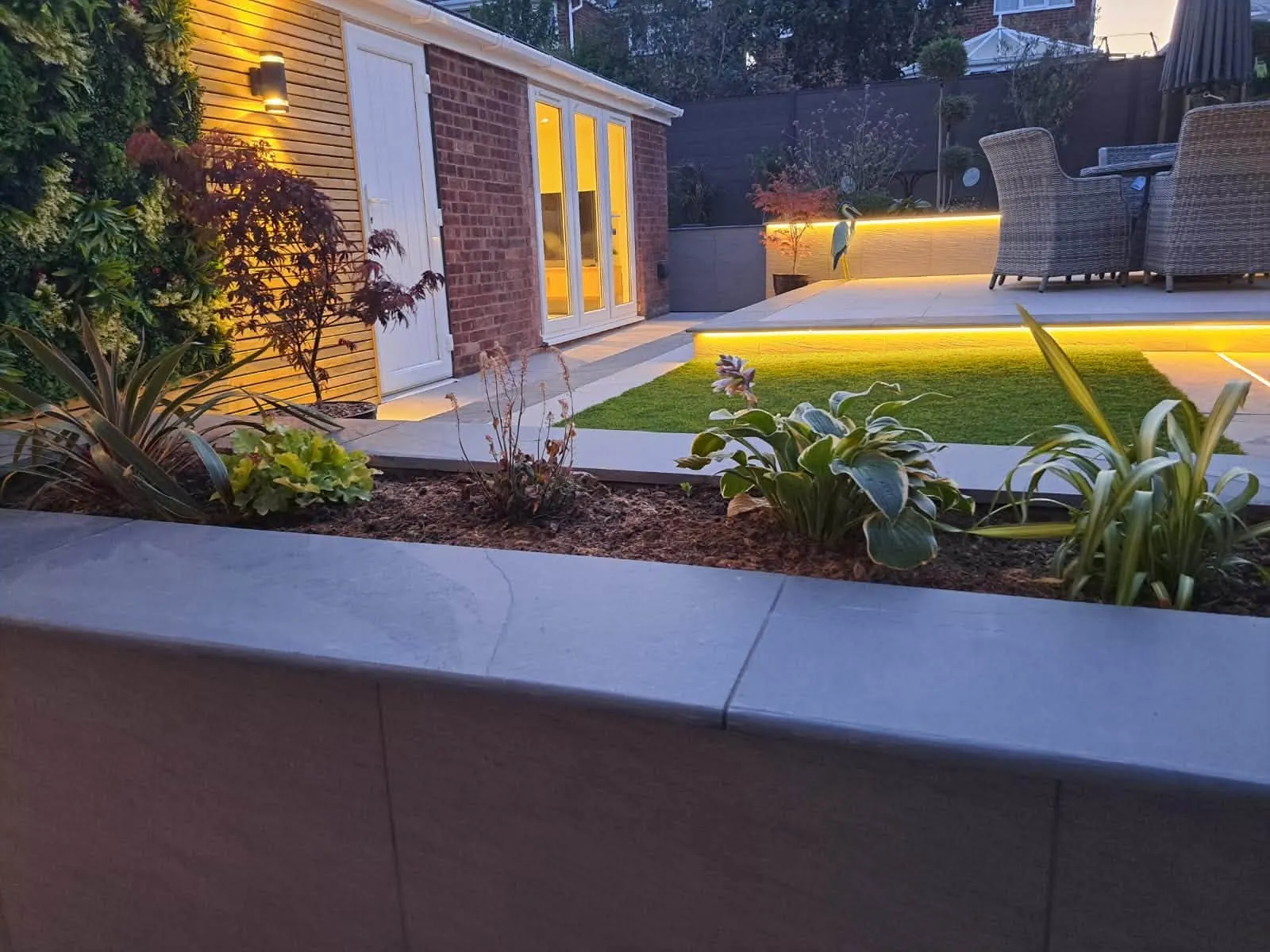 Garden with illuminated raised patio, lawn and planting beds at dusk