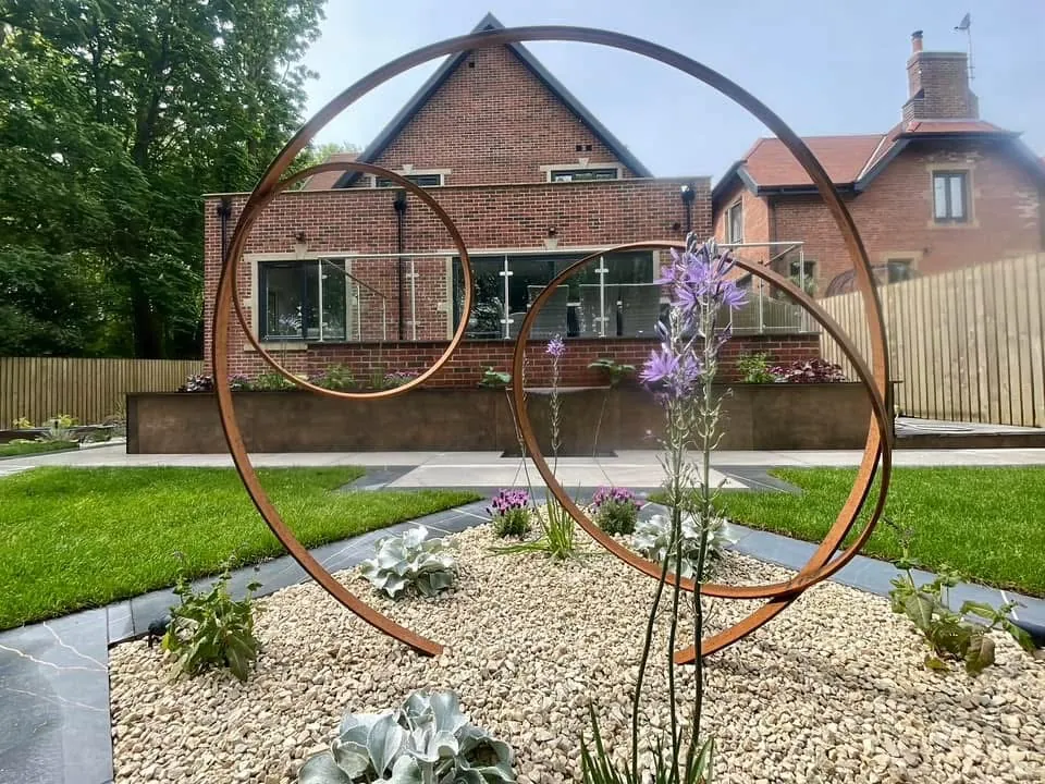Corten steel ring sculpture in a gravel border with lawn and paving